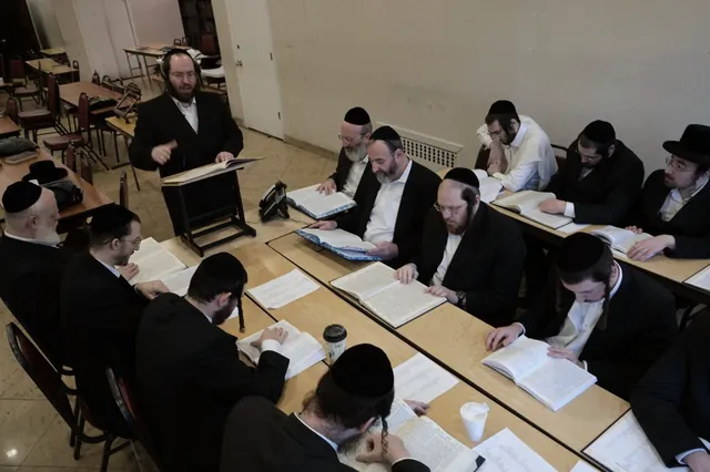 א בליק פון אויבן אויף די טישן מיט אפענע גמרות און צוהערער, און רבי משה אליעזר שליט״א ביים שטענדער אין עק פונעם צימער