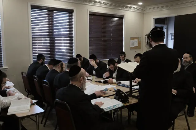 א צימער פול מיט צוהערער וואס לערנען ביי די טישן, און רבי משה אליעזר שליט״א שטייט ביים שטענדער מיטן רוקן צום אפאראט